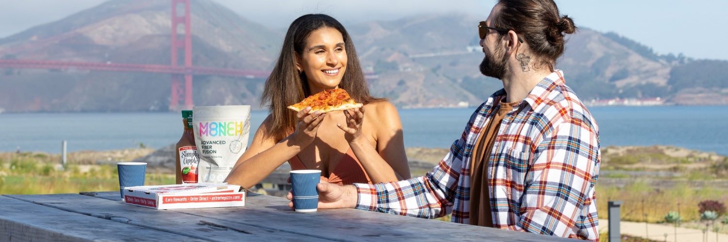 A couple sitting at a picnic table outdoors, smiling and enjoying pizza. A bag of Monch Monch original product is positioned in front of them