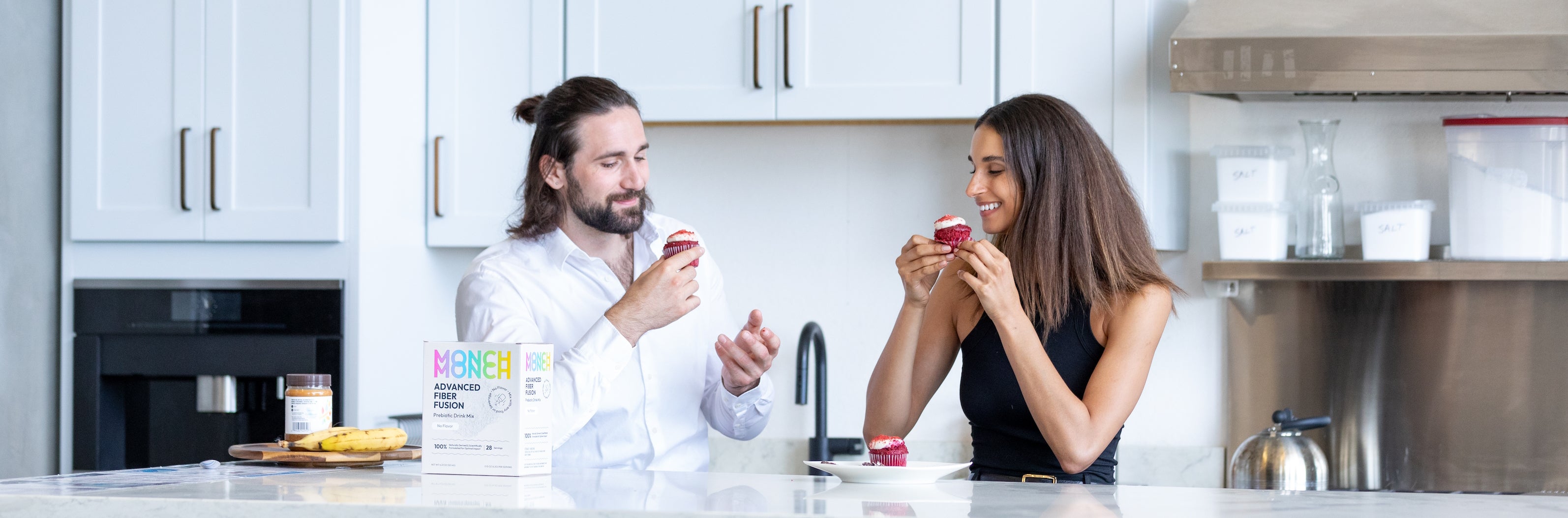 A couple sitting at a kitchen counter, smiling and enjoying cupcakes. A box of Monch Monch original product is positioned in front of them