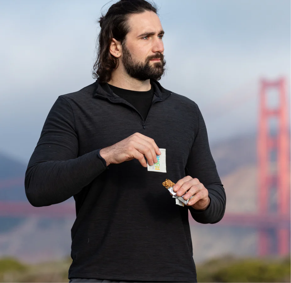 Man holding Monch Monch Original sachet and a granola bar while outdoors, with the golden gate bridge in the distant background