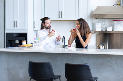 A couple sitting at a kitchen counter, smiling and enjoying cupcakes. A box of Monch Monch original product is positioned in front of them