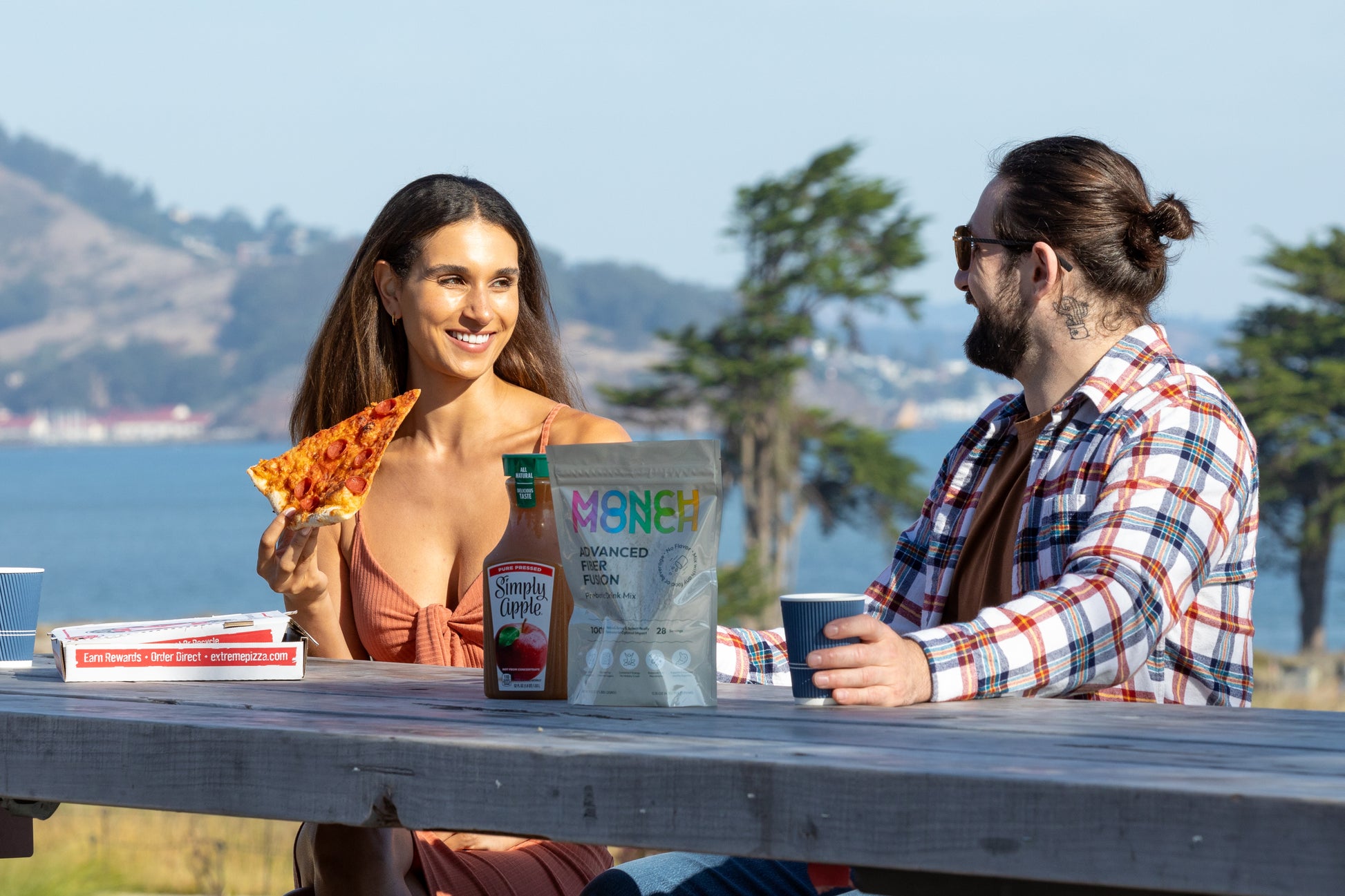 A couple sitting at a picnic table outdoors, smiling and enjoying pizza. A bag of Monch Monch original product is positioned in front of them